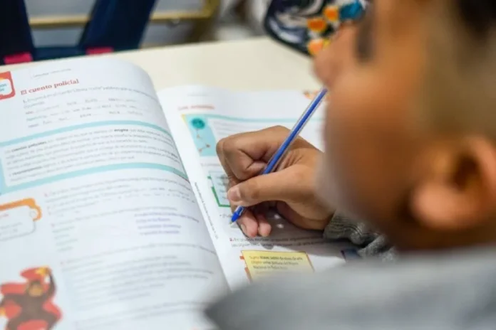 Niños realizando tareas escolares en una mesa, acompañados por un docente, en un espacio comunitario.
